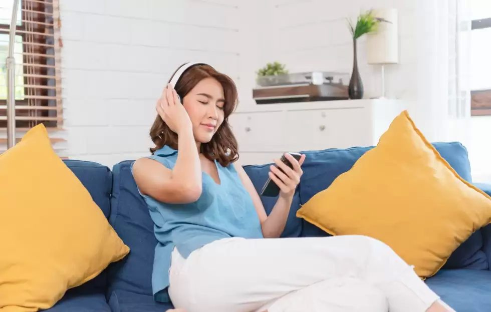 young-asian-woman-using-smartphone-listening-music-by-connecting-headphone-sitting-sofa-living-room-weekend