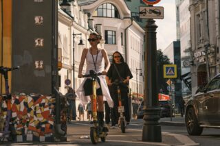 Two women riding electric scooters on a city street, showcasing urban commute.