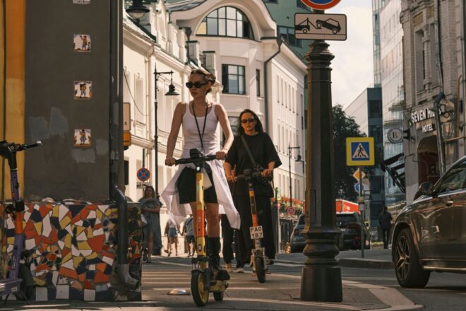 Two women riding electric scooters on a city street, showcasing urban commute.